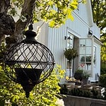 En svart, sfärisk ampel vid namn Ragna från varumärket Wikholm form, hänger i ett träd med blommande grenar i en grönskande trädgård. I bakgrunden syns en vit byggnad med öppna dörrar och fönster, omgiven av blommande växter och buskar.