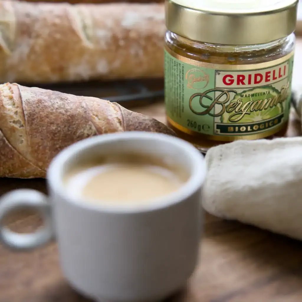 En burk Bergamottmarmelad från Gridelli, omgiven av nybakat bröd och en kopp kaffe, placerad på ett träbord.