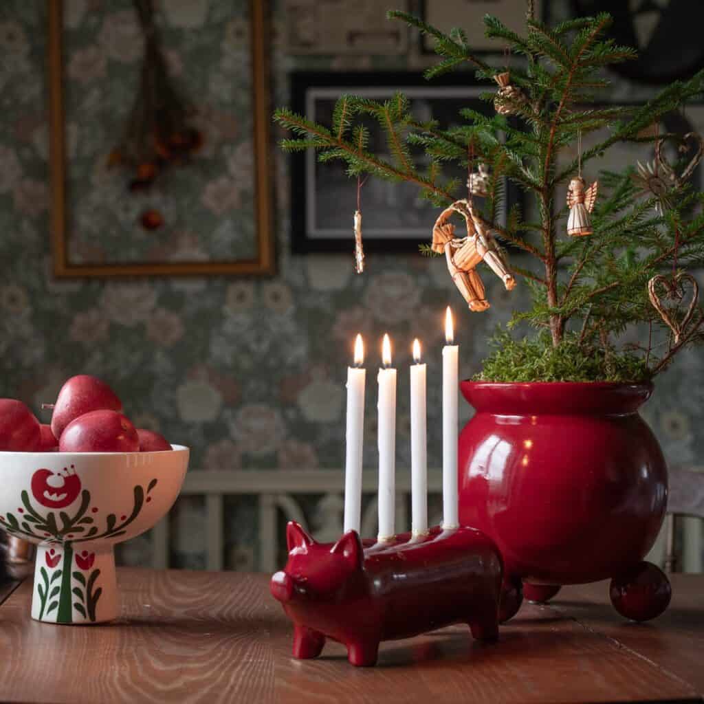 Juldekoration med kruka ANYA röd med en liten gran och halmänglar, tillsammans med en ljusstake och en skål med röda äpplen på ett träbord.