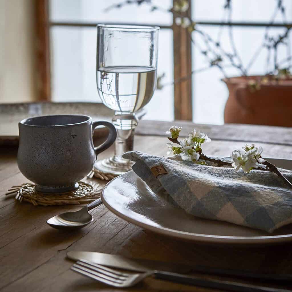 En dukning med en beige tallrik från serien Annabelle av Affari of Sweden. På tallriken ligger en blå- och vitrutiga servett dekorerad med en kvist med vita blommor. Vid sidan av tallriken finns en kopp och ett glas med vatten, båda placerade på flätade underlägg. I bakgrunden syns en blommande växt vid ett fönster.