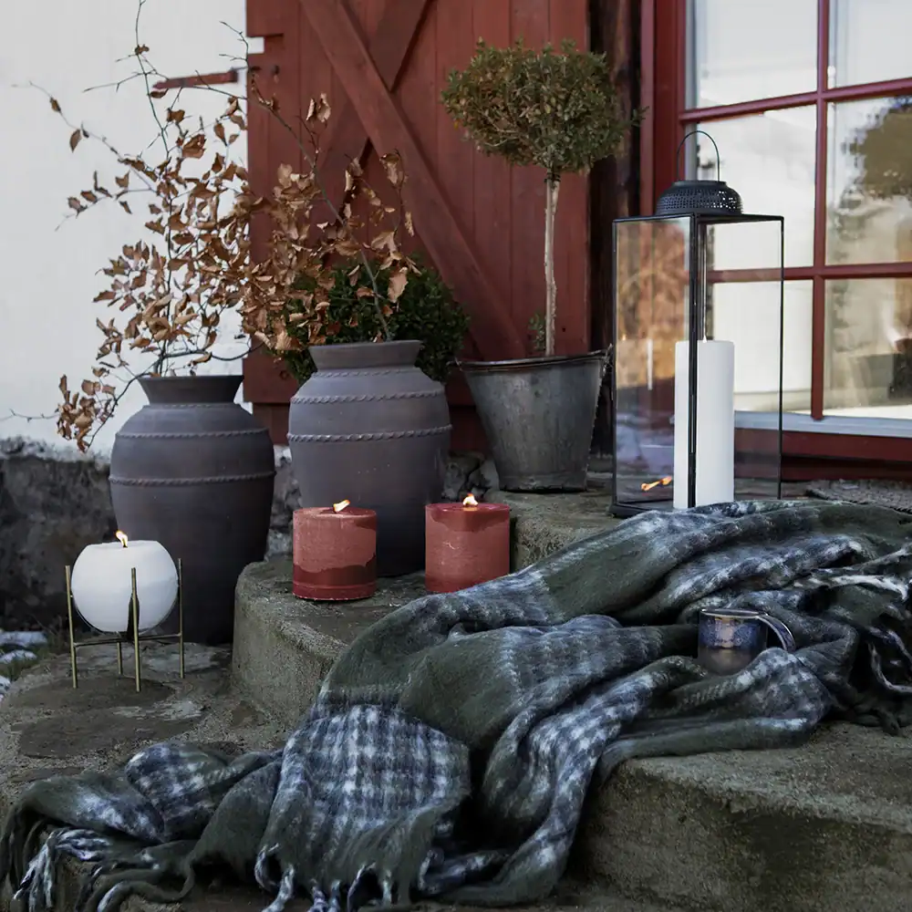 En stämningsfull höstscen utomhus med röda och vita ljus från Affari of Sweden som brinner på stentrappan. I bakgrunden syns en träddörr och fönster, omgivna av vaser och trädgårdskrukor mot en vit vägg. En mysig filt ligger draperad över trappan, vilket skapar en inbjudande och varm atmosfär.