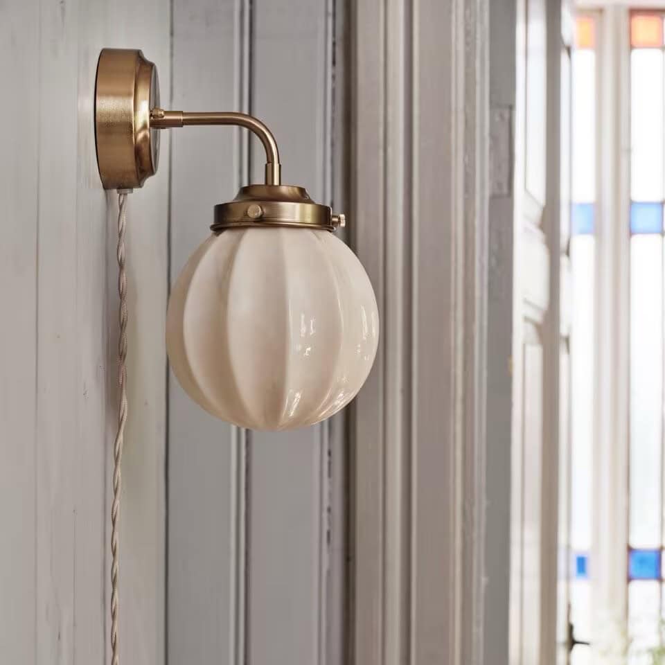 Vägglampan Marit från Strömshaga på en grå trävägg i ett rum med färgade fönster. Lampans opalglas reflekterar ljuset i rummet.