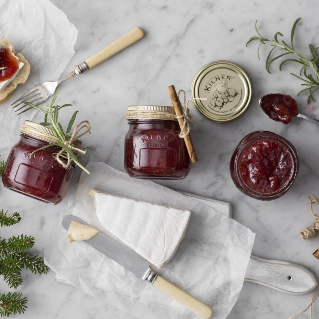 Flera Kilner konserveringsburkar fyllda med röd marmelad, dekorerade med kanelstänger och rosmarin, tillsammans med en ostbricka.