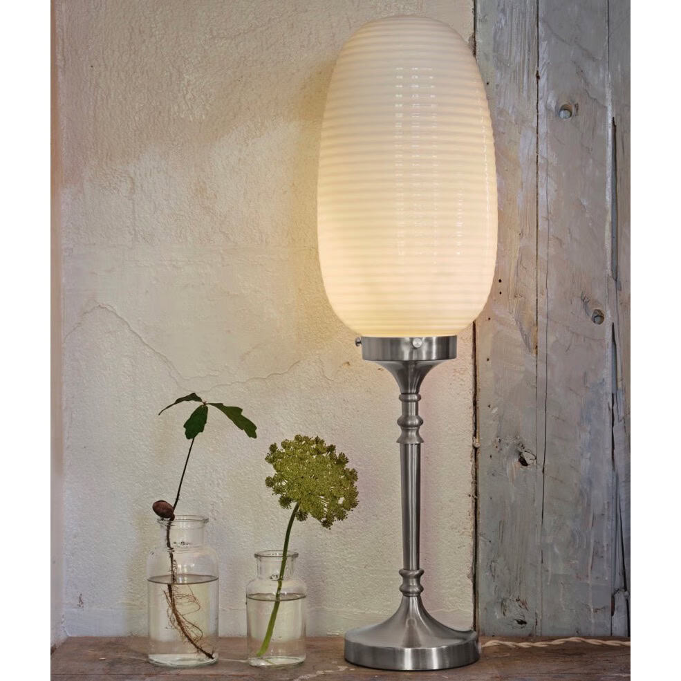 Bordslampa Evald från Strömshaga med avlång glaskupa i frostat vitt och en dekorativ fot i förnicklad metall, placerad mot en rustik bakgrund med glasvaser och gröna växter.