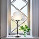 Bordslampa Eli från Strömshaga placerad i ett fönster, med frostad glaskupa och fot i förnicklad metall som sprider ett varmt ljus.