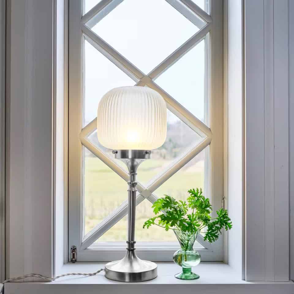 Bordslampa Eli från Strömshaga placerad i ett fönster, med frostad glaskupa och fot i förnicklad metall som sprider ett varmt ljus.