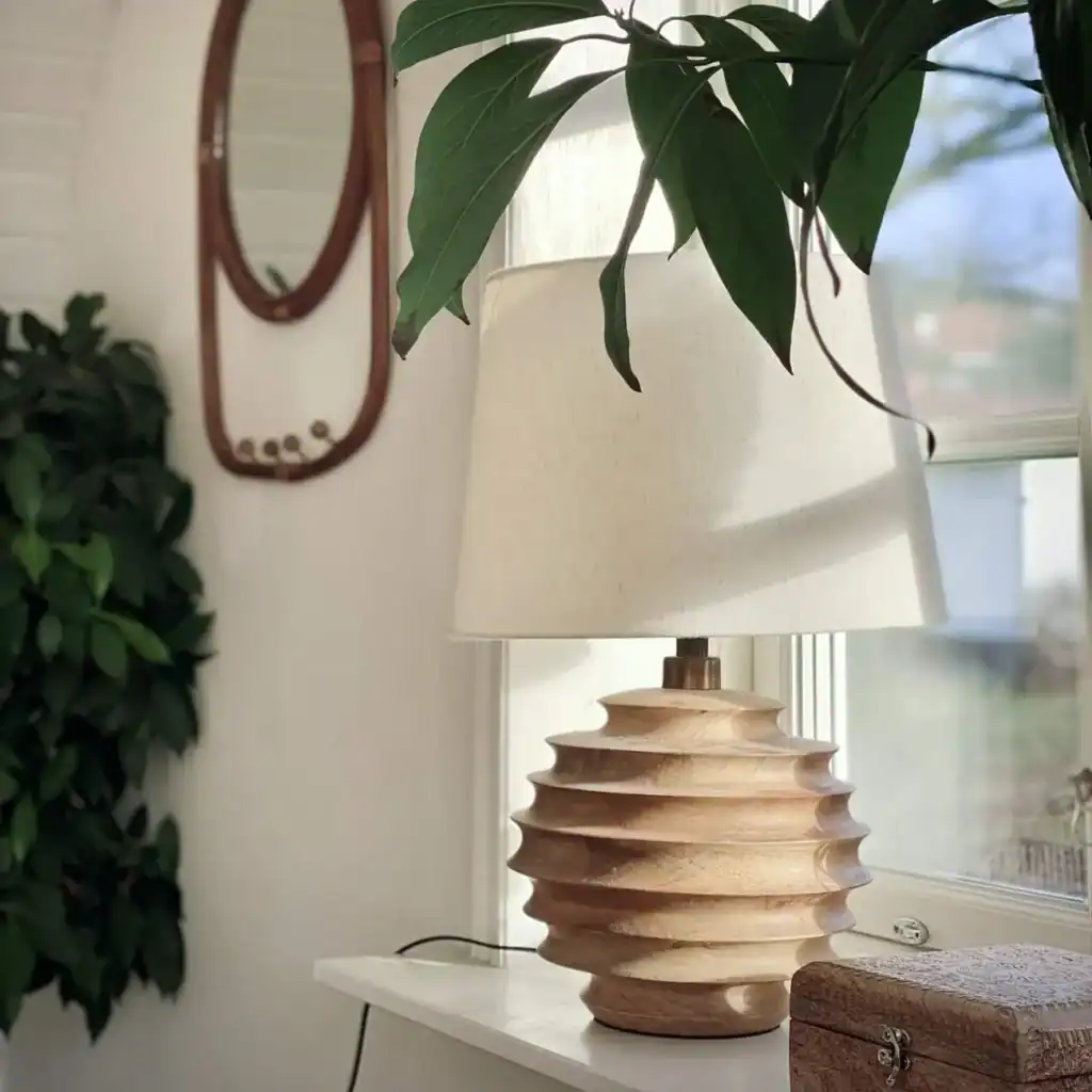 Bordslampa med en design i ljust trä och en vit lampskärm står på ett fönsterbräde. Omringad av gröna växter och detaljer i naturmaterial, ger den inspiration för inredningstips för hemmet.
