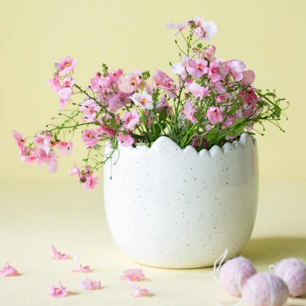Äggformad kruka Mona i vitmelerad design, fylld med rosa blommor och stylad mot en gul bakgrund, perfekt för påskdekoration och vårens inredning.