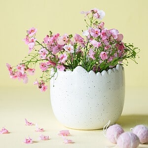 Äggformad kruka Mona i vitmelerad design, fylld med rosa blommor och stylad mot en gul bakgrund, perfekt för påskdekoration och vårens inredning.