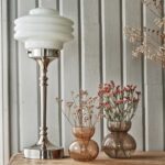 Bordslampa Valter från Strömshaga i förnicklad metall och vit opalglas-kupa, placerad på ett träbord bredvid bruna glasvaser fyllda med torkade och rosa blommor, mot en grå träpanelvägg.