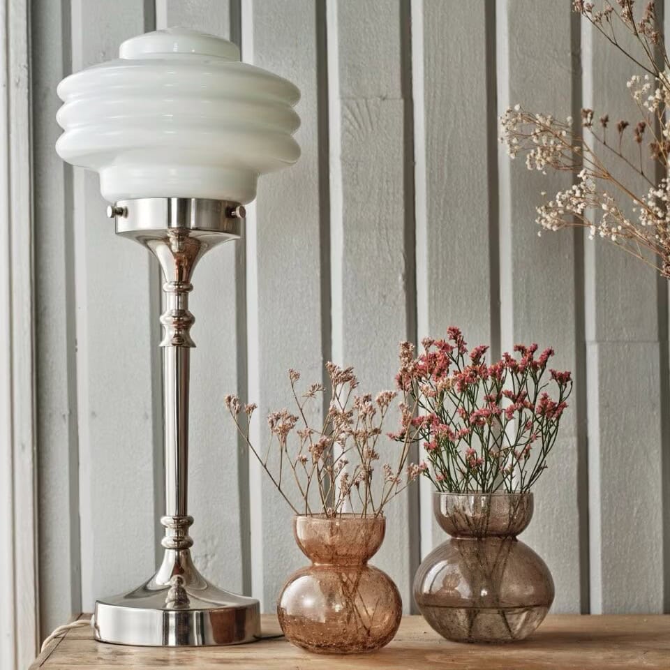 Bordslampa Valter från Strömshaga i förnicklad metall och vit opalglas-kupa, placerad på ett träbord bredvid bruna glasvaser fyllda med torkade och rosa blommor, mot en grå träpanelvägg.