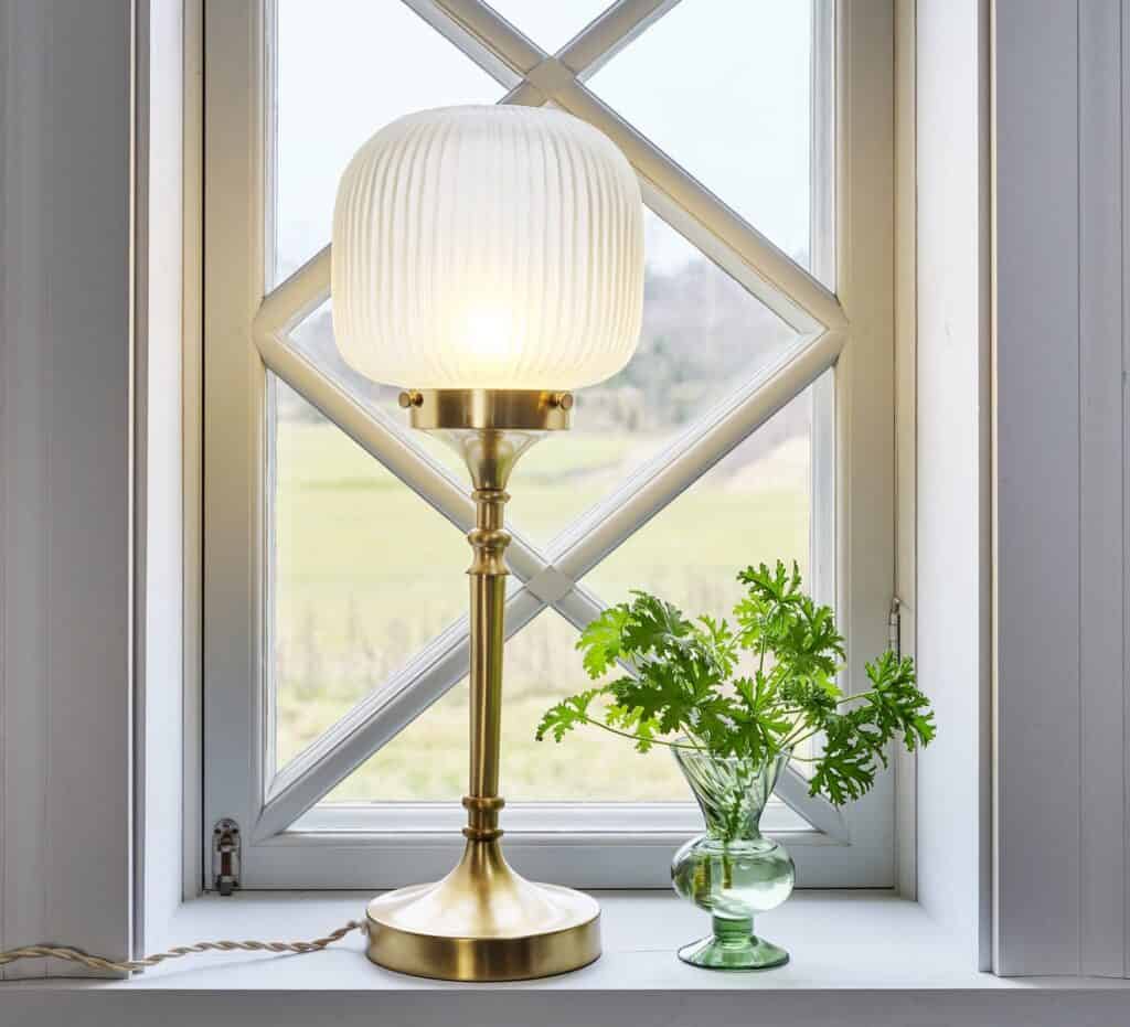 Elegant bordslampa i mässing med vit veckad glasskärm, placerad på ett fönsterbräde tillsammans med en grön växt i en glasvas – ett stilfullt exempel på inredningstips för hemmet.