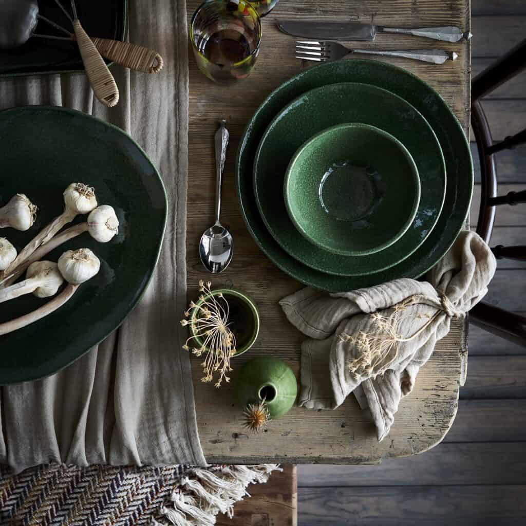 Rustik dukning med gröna tallrikar och skålar från ANNABELLE-kollektionen på ett träbord, omgiven av linneservetter, vitlök och keramikdetaljer.