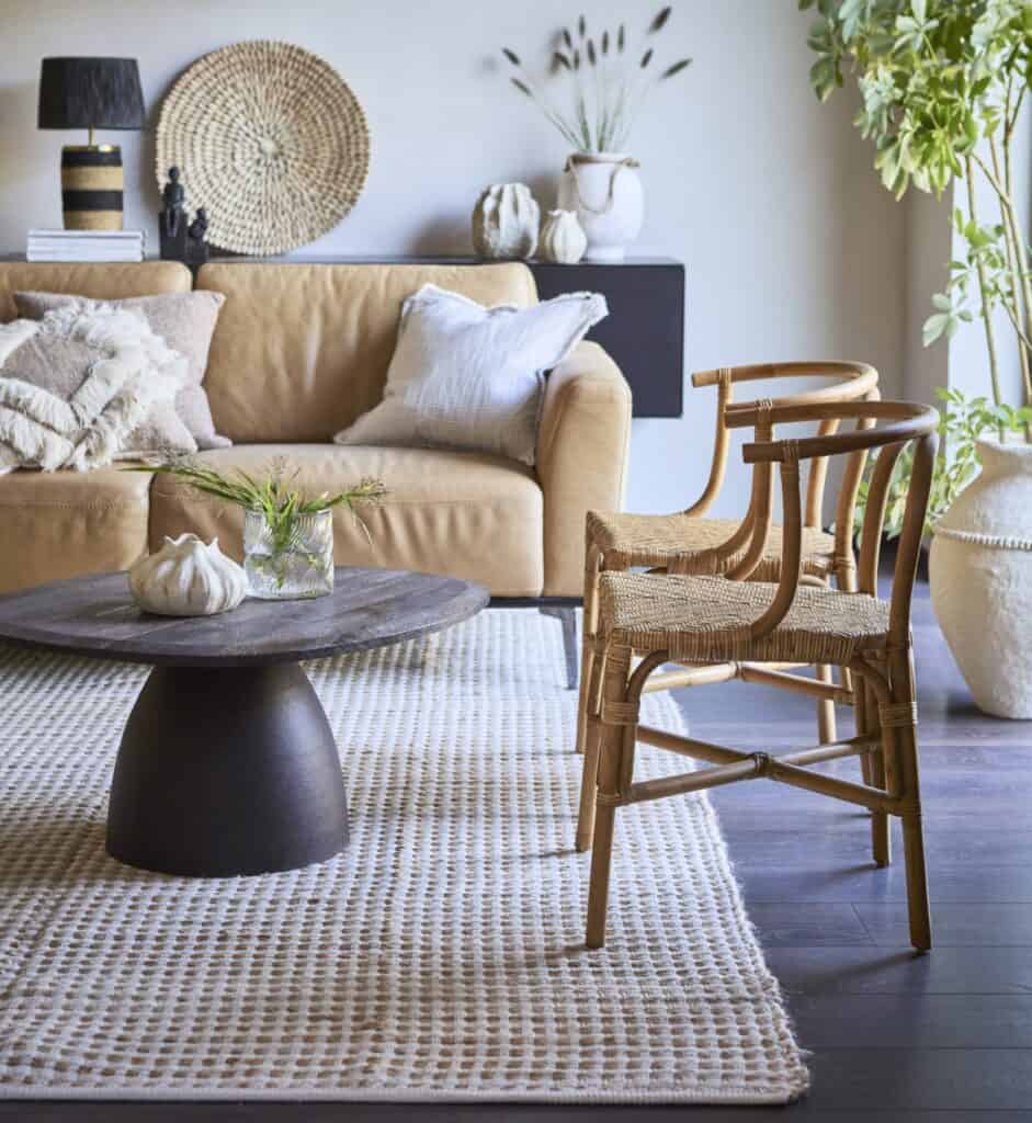 Mysigt vardagsrum med en beige soffa, mjuka kuddar, en ljus matta och naturinspirerade detaljer som rottingstolar och gröna växter – ett inspirerande exempel på hur textilier kan förnya inredningen.