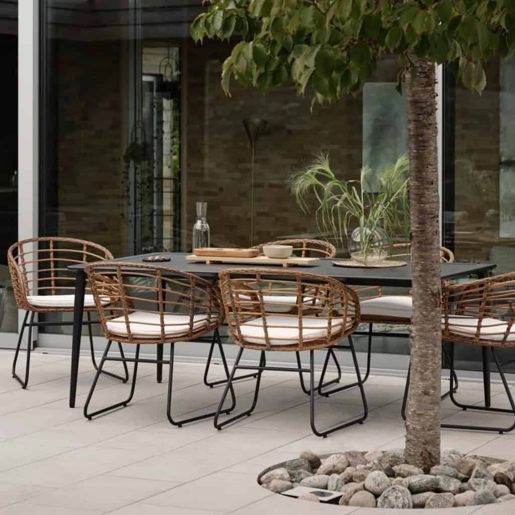 Utemiljö med matgruppen Covelo från Brafab. Stilrena matstolar i naturfärgad konstrotting med svart metallram, placerade runt ett modernt svart matbord på en elegant terrass.