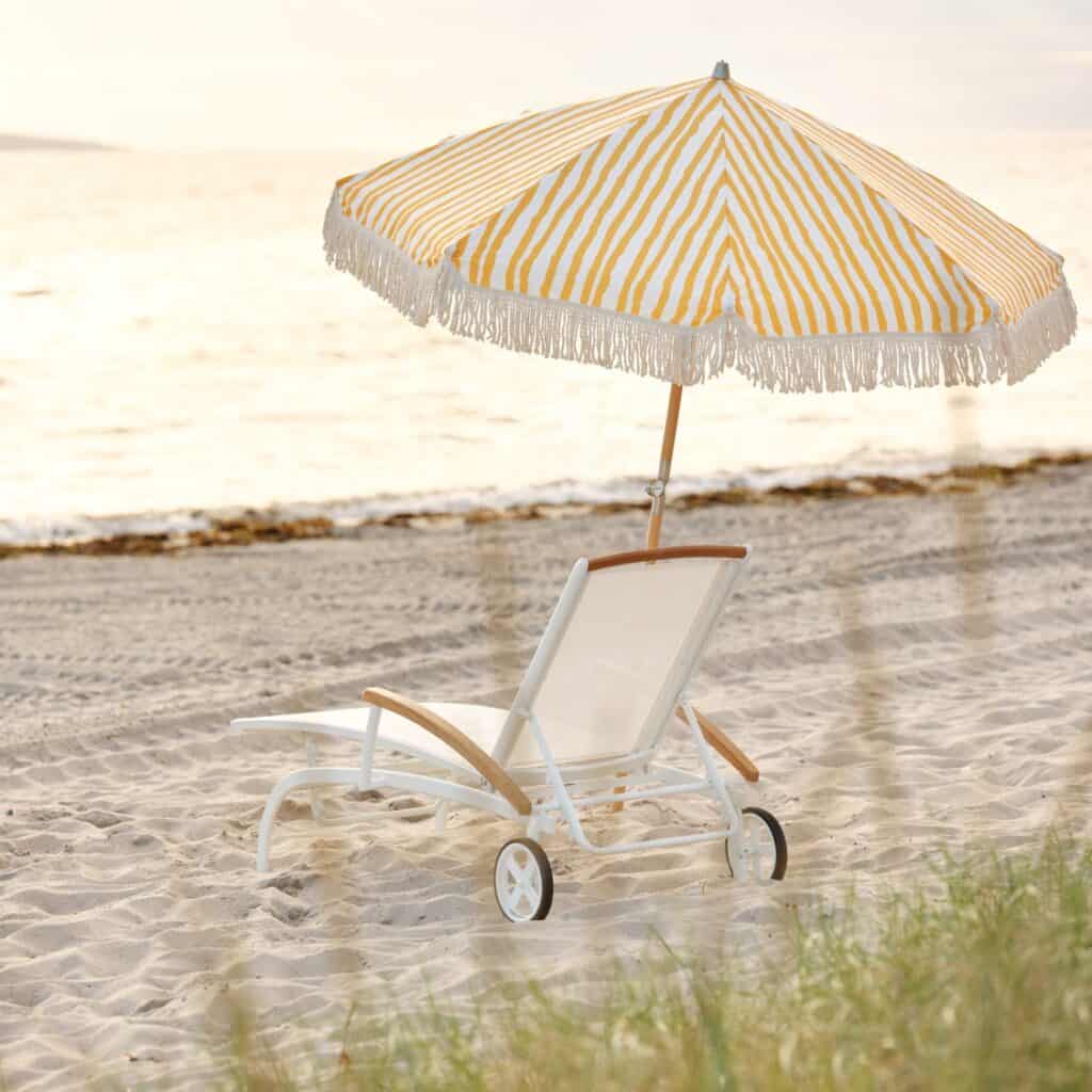 Gatsby parasoll i gul- och vitrandigt mönster med fransar, uppsatt vid en solstol på en sandstrand vid havet i solnedgången.