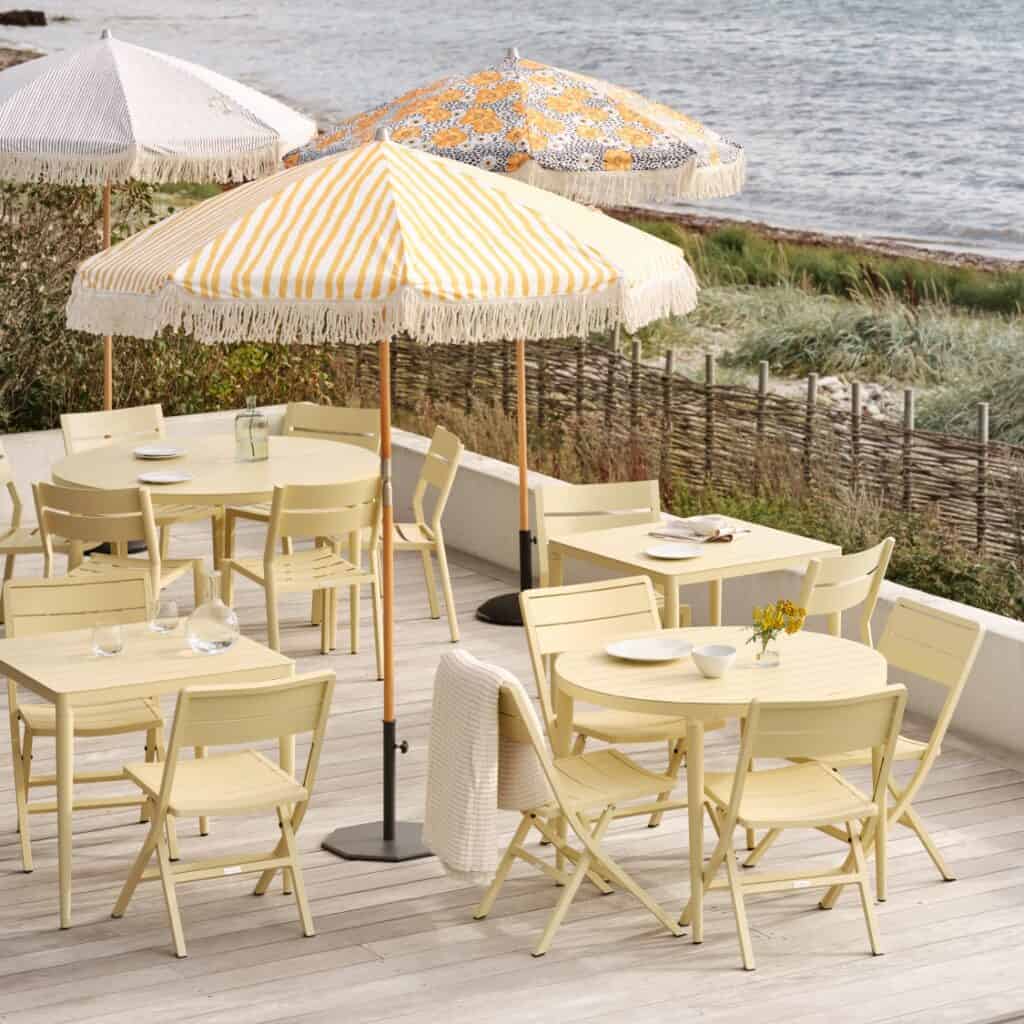 Uteservering med Gatsby parasoll från Brafab i gul- och vitrandigt mönster med fransar, omgiven av ljusgula trädgårdsmöbler vid havet.