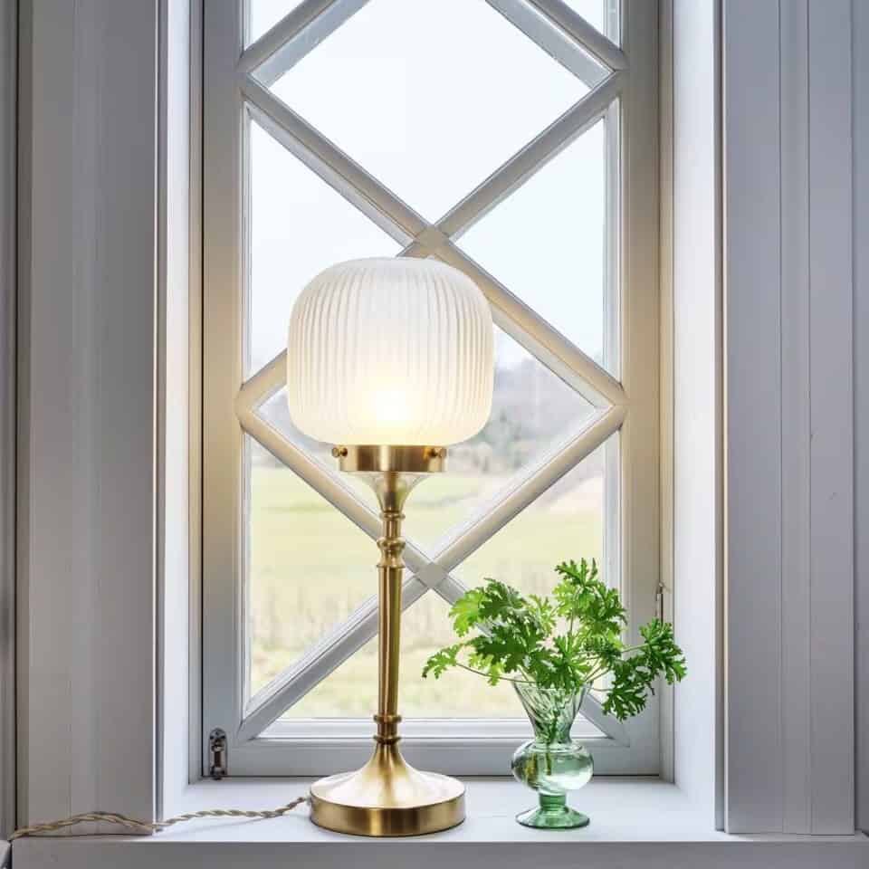 Bordslampa med frostat glas och mässingsfot – Strömshaga Eli i fönster med grön växt