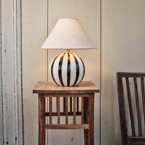 Lampfot ALBA från Strömshaga med svartvit randig glasfot och beige lampskärmen Hedvig, placerad på ett rustikt träbord mot en ljus trävägg.