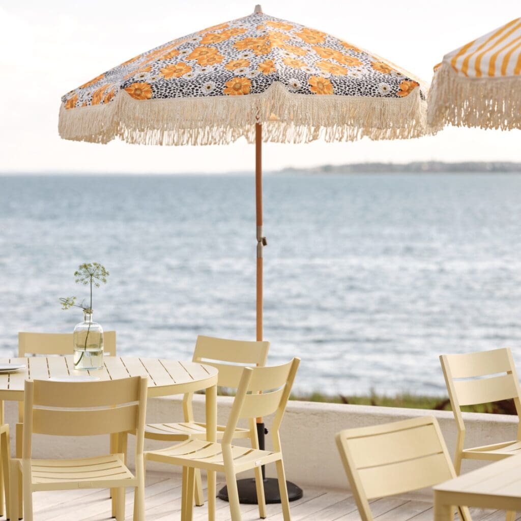 Parasoll Gatsby blommig 185 cm från Brafab med franskant, uppställt på altan vid havet tillsammans med ljusgula utemöbler.