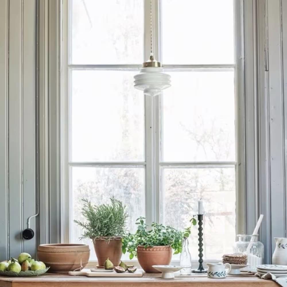 Taklampa med tvinnad sladd i mässing och vit opalglas från Strömshaga, upphängd framför ett fönster i lantligt kök.