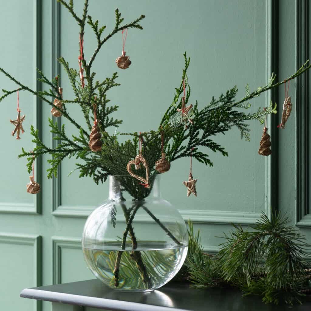 Halmdekorationer till jul hängande på grankvist i glasvas – naturlig julinredning i skandinavisk stil.