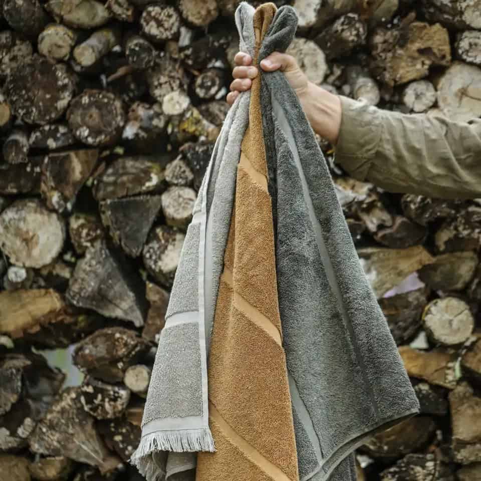 FIGARO handdukar i beige, rökt ek och grönt hålls framför vedstapel