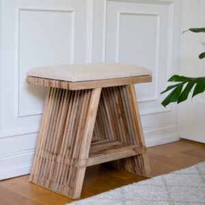 Pall i teak med beige tygsits i hemmiljö framför vit vägg, användbar som extra sittplats, sidobord eller dekorativ möbel.